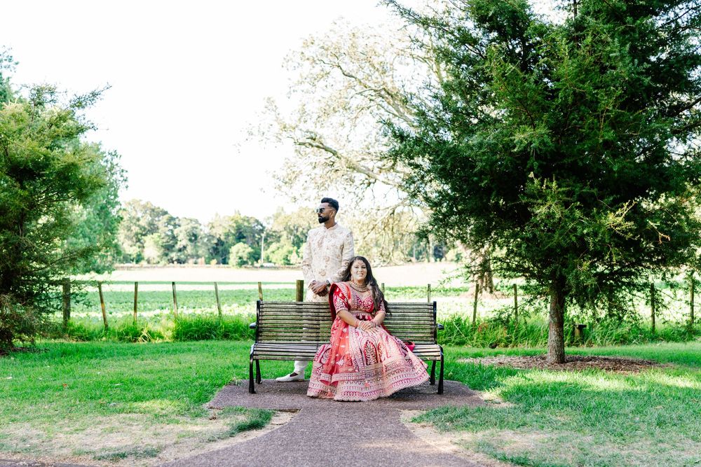 Two people sit on a wooden bench in a scenic garden setting with evergreen trees and sunlit meadow in background.
