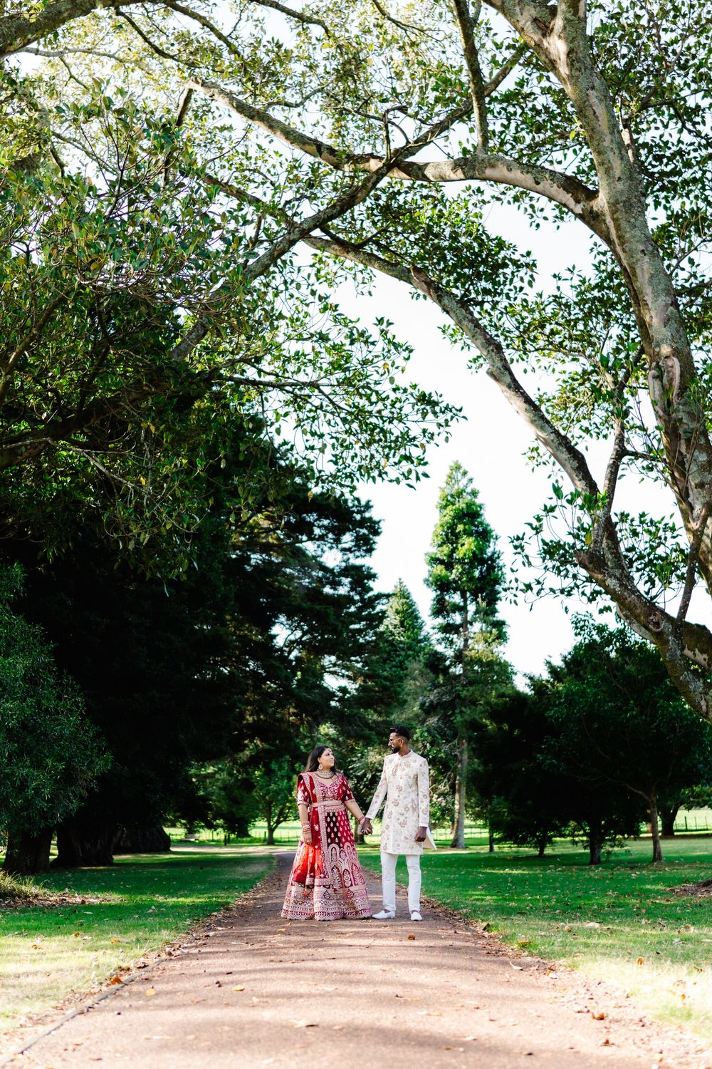A couple takes a romantic stroll down a tree-lined dirt path in a lush green park setting.