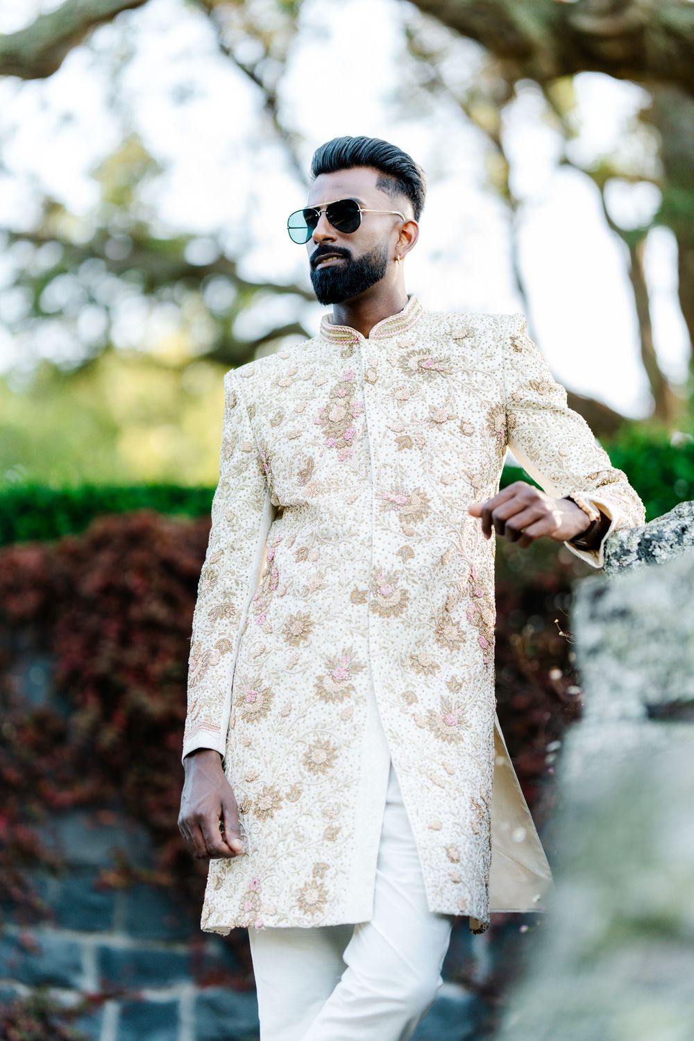 A person wearing sunglasses and a cream-colored floral print jacket poses against a blurred outdoor background.