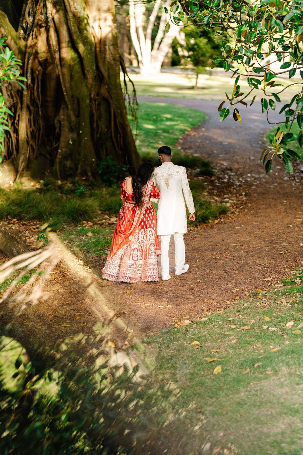 A couple in traditional Indian wedding attire walk together on a shaded garden path surrounded by lush greenery.
