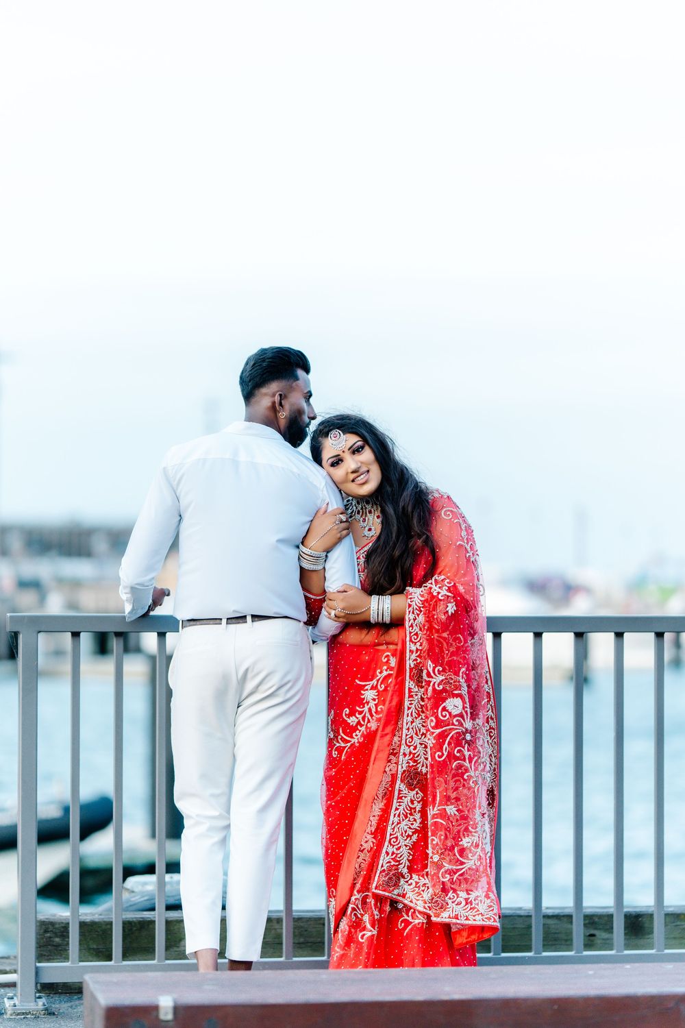 A couple in formal attire, with a red saree, embraces on a balcony overlooking a cityscape.