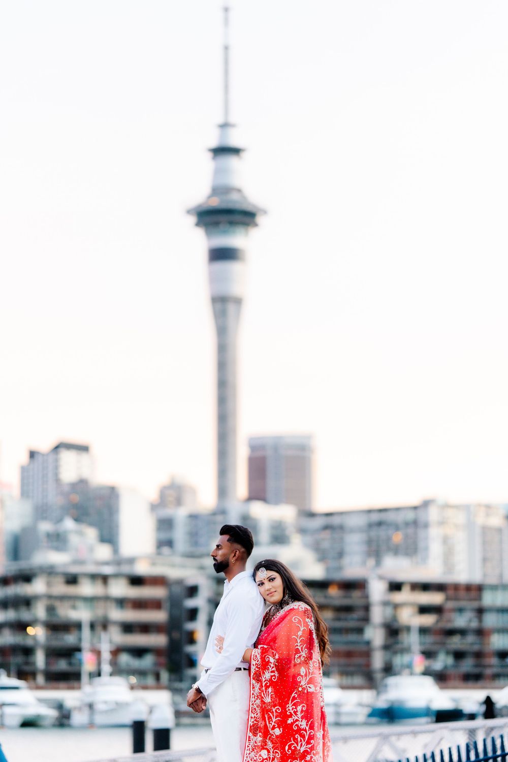 Couple in romantic outfits poses along city waterfront with skyline view and Sky Tower in background.