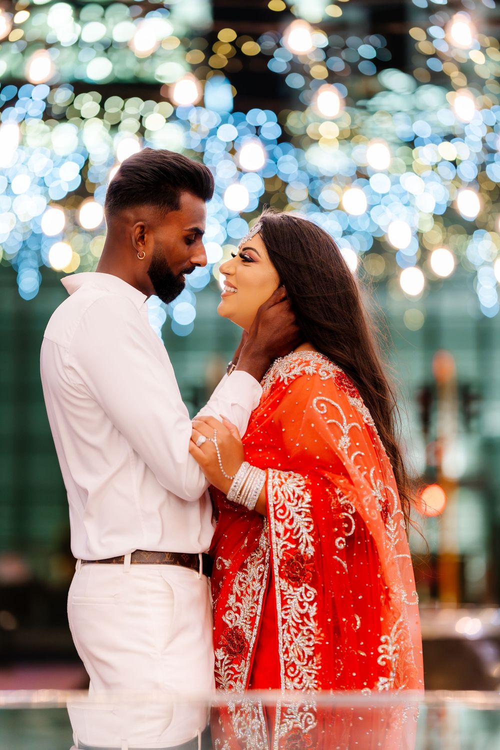A couple shares a romantic moment under twinkling bokeh lights, with one wearing white and the other in a red sari.