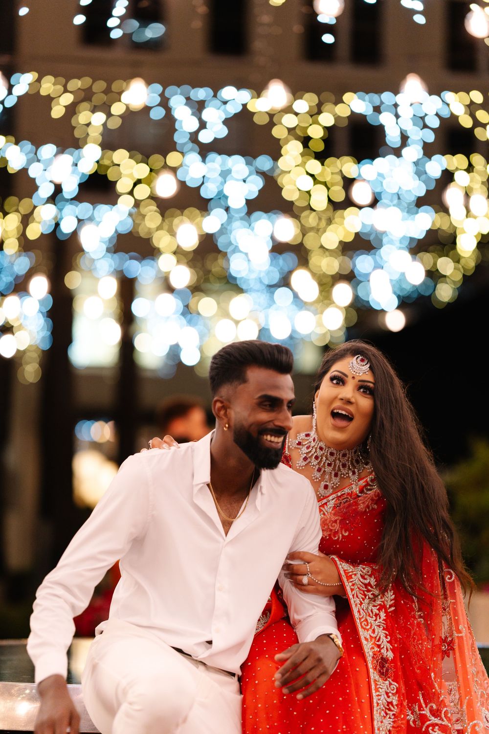 Two people sharing a joyful moment under twinkling string lights, with one wearing an elegant red and gold traditional outfit.