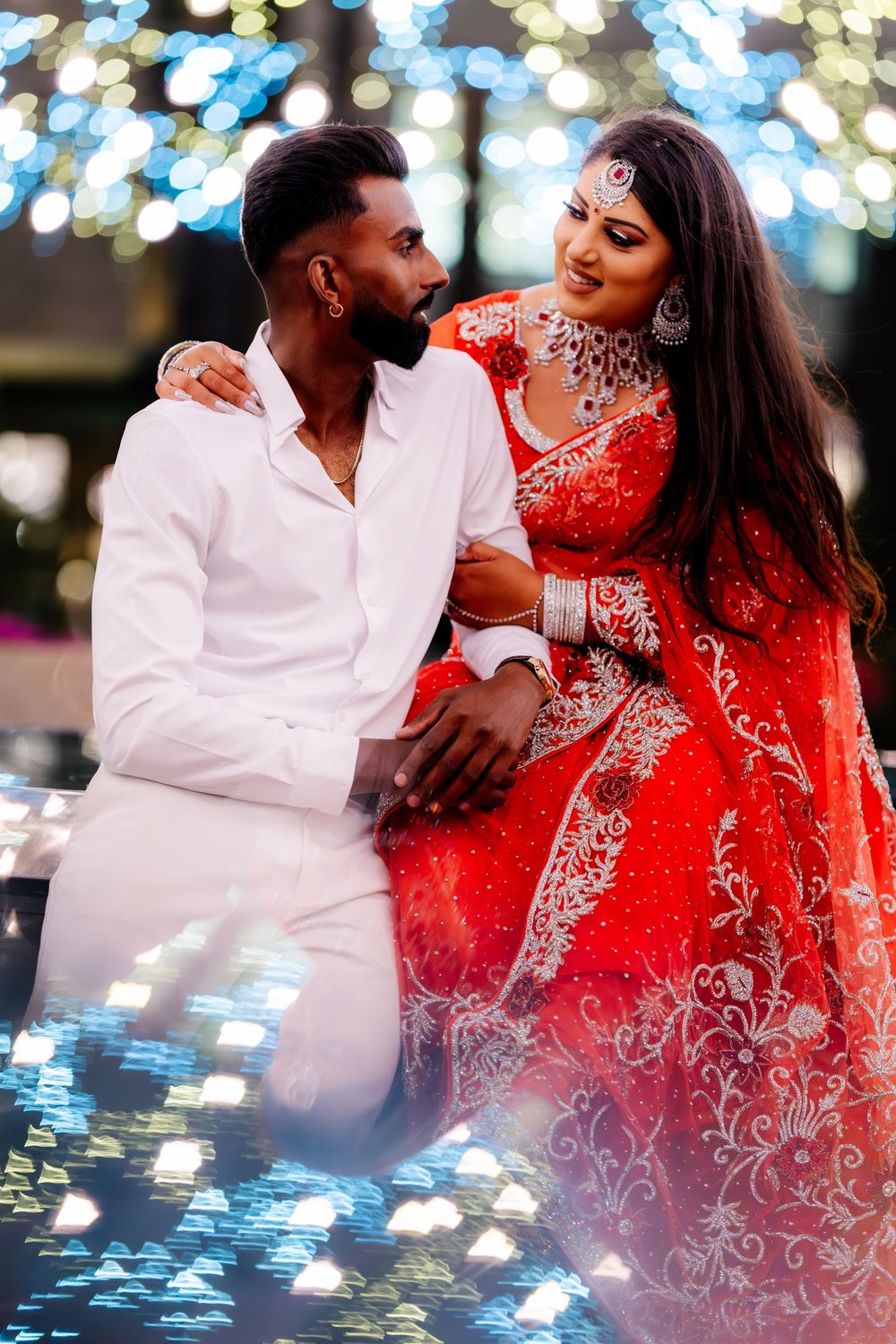 An elegant couple embraces in traditional wedding attire featuring a red embellished dress against bokeh lights.
