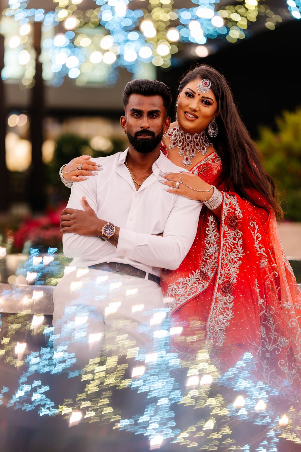 A romantic couple poses together outdoors wearing traditional Indian wedding attire with glowing bokeh lights in background.