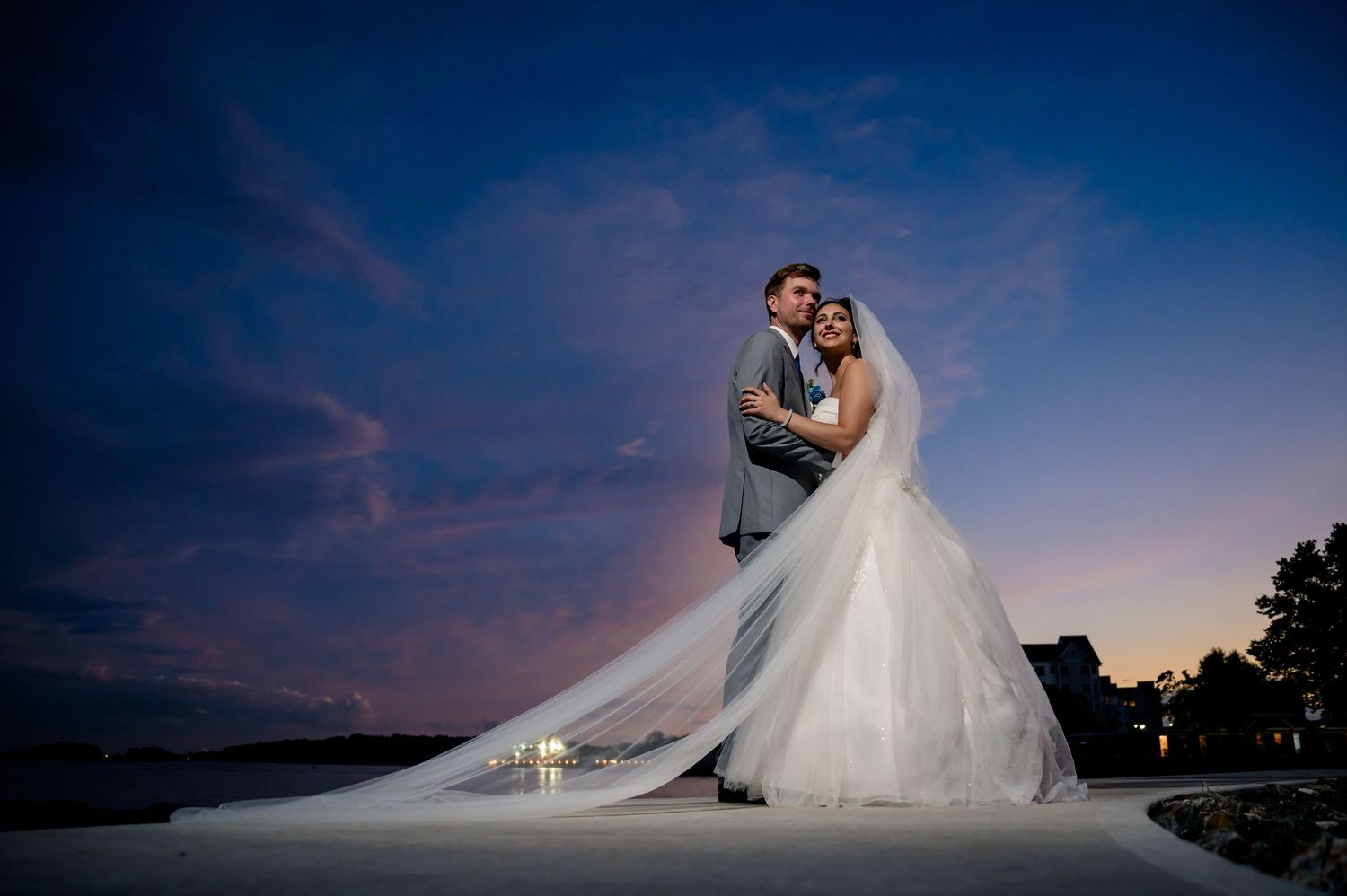 A couple in wedding attire embrace against a dramatic twilight sky as the bride's veil flows in the wind.