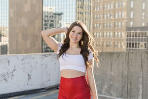 Model in red skirt and white crop top poses confidently against urban backdrop near brick and stone walls.