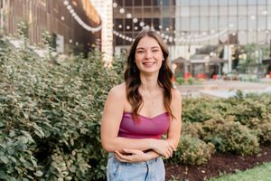 A person in a pink top and jeans smiles while standing in front of landscaping and string lights.