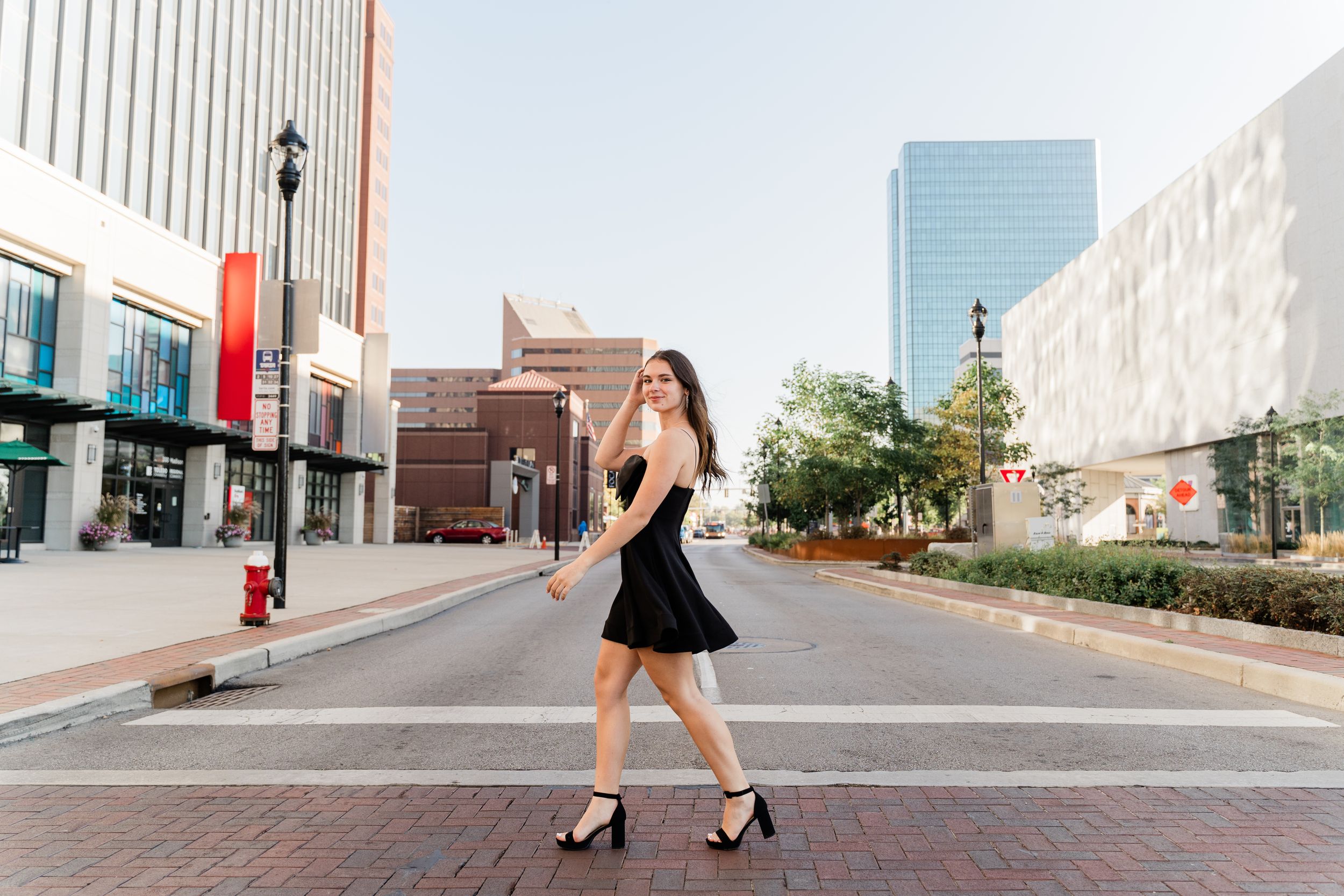 A person in a black dress and heels poses on a downtown street with modern buildings and lamp posts in the background.