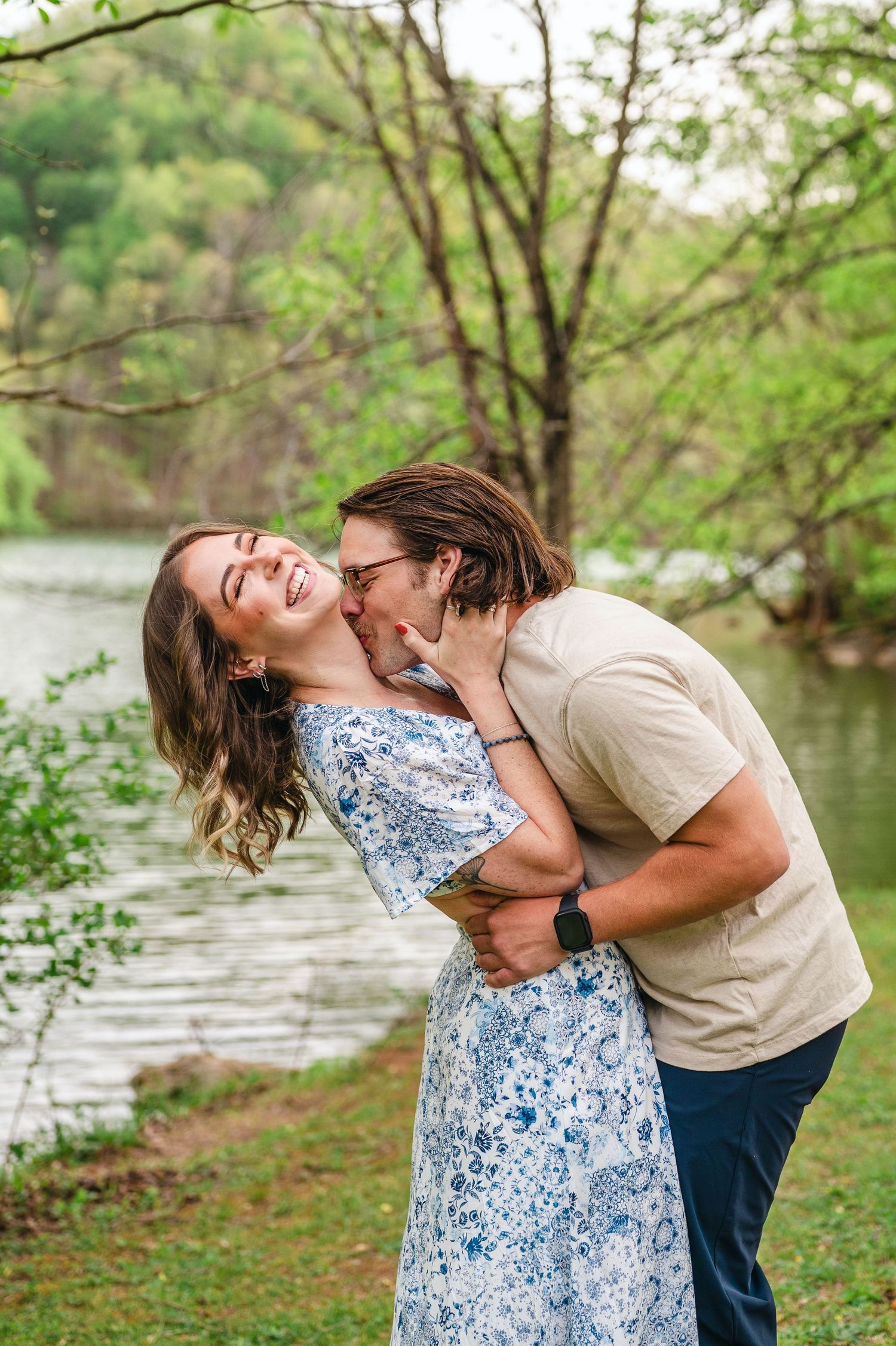 A romantic moment captured by a North Carolina lake with spring trees, featuring a blue floral dress and casual attire.