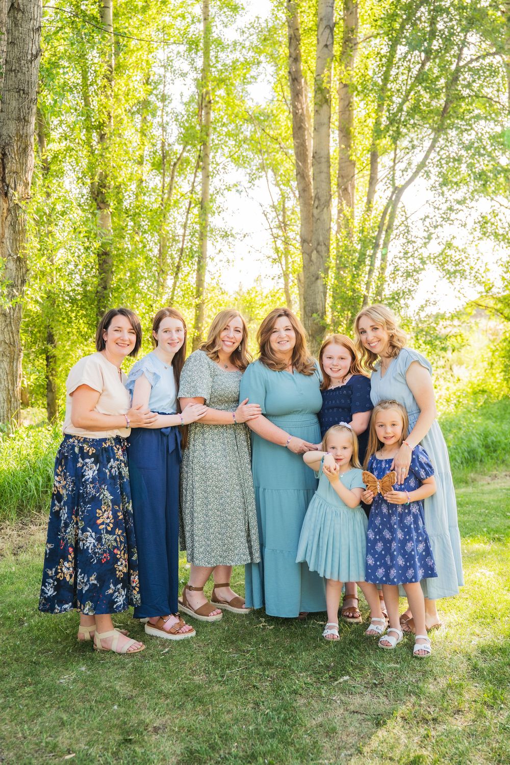 A group of people in coordinated blue and floral dresses pose together on a sunny day in a wooded outdoor setting.