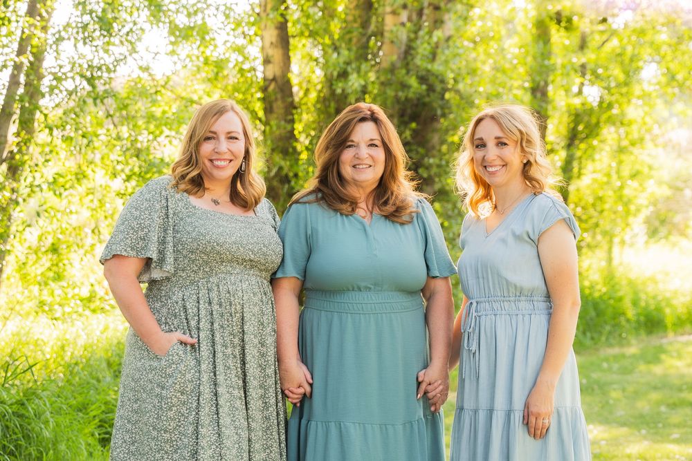 Three women in coordinating sage and teal colored midi dresses stand together in a sunny outdoor setting.