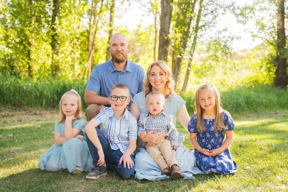 A cheerful family sits together on grass in a sunlit wooded setting during a summer photo session.
