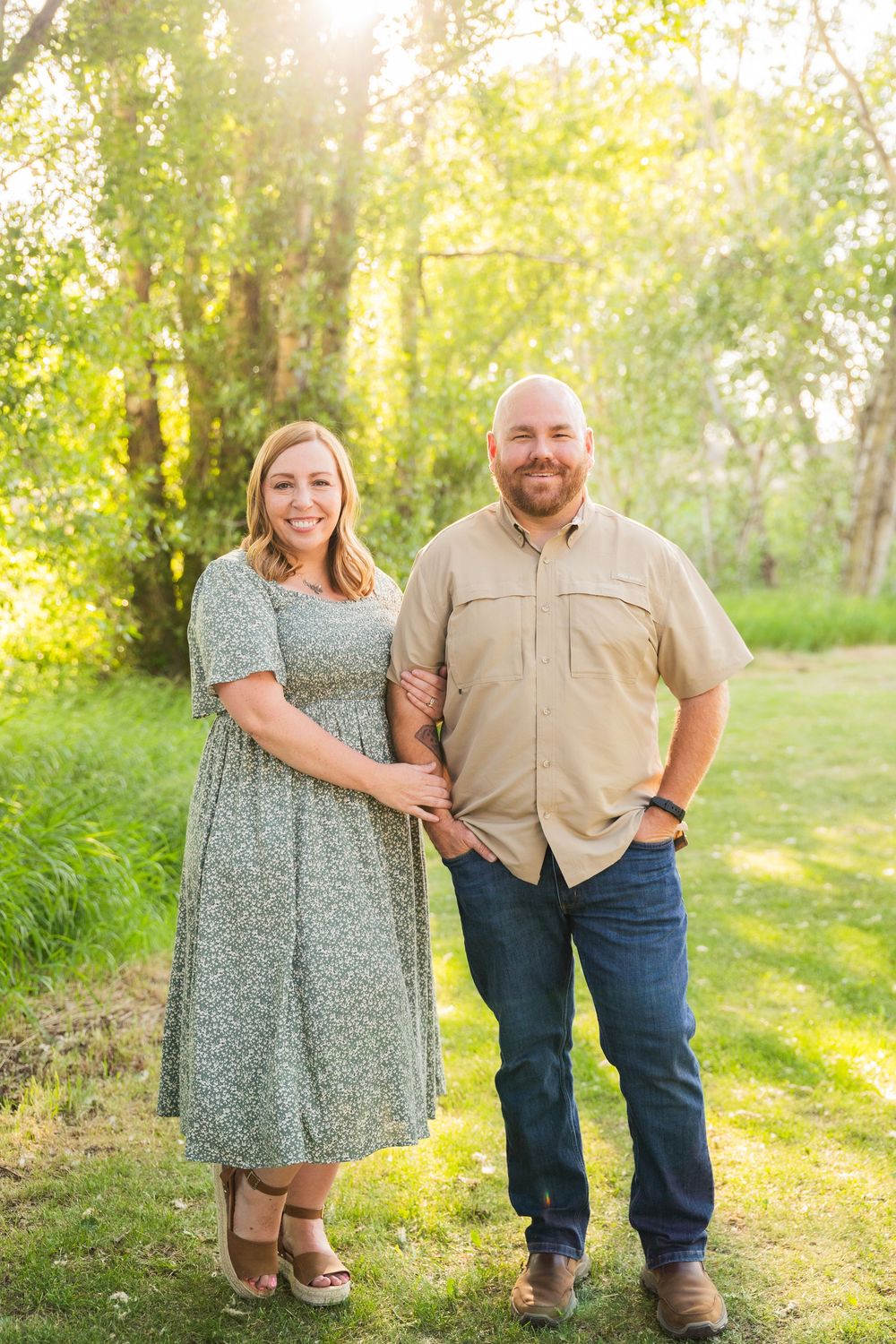 A couple stands in a sunlit grassy meadow wearing coordinated casual outfits during a romantic outdoor photoshoot.