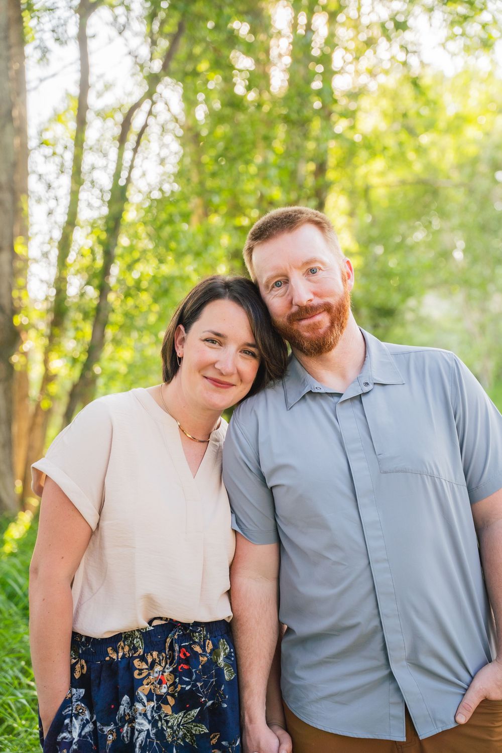 A couple poses together outdoors in a sunlit wooded setting with green foliage in the background.
