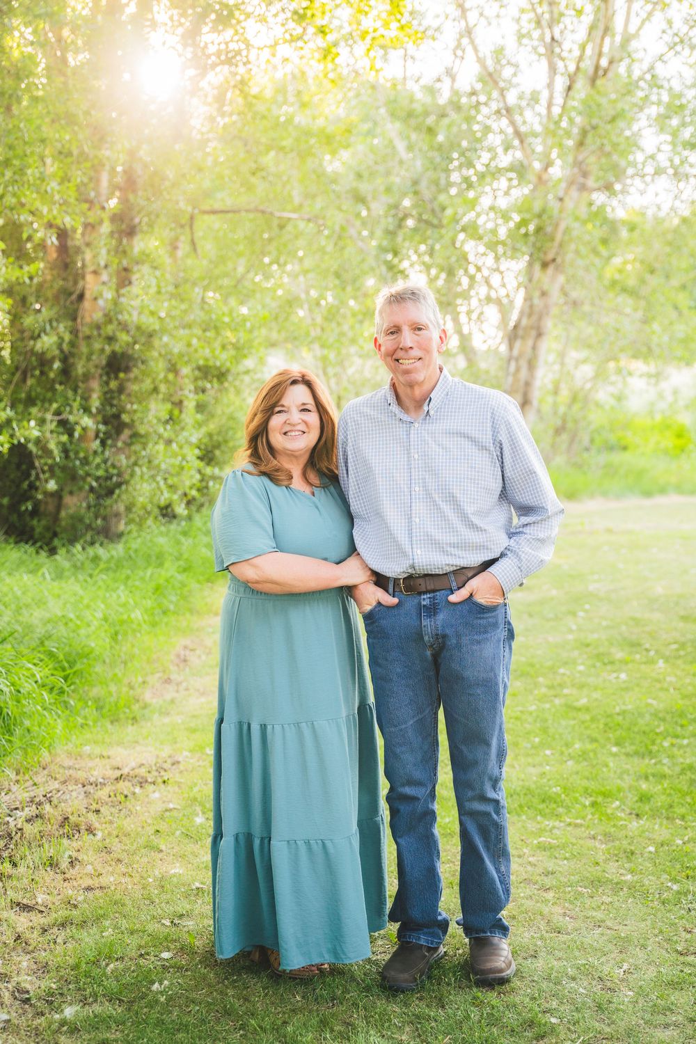 A couple poses together outdoors on a sunny day with trees and greenery in the background.