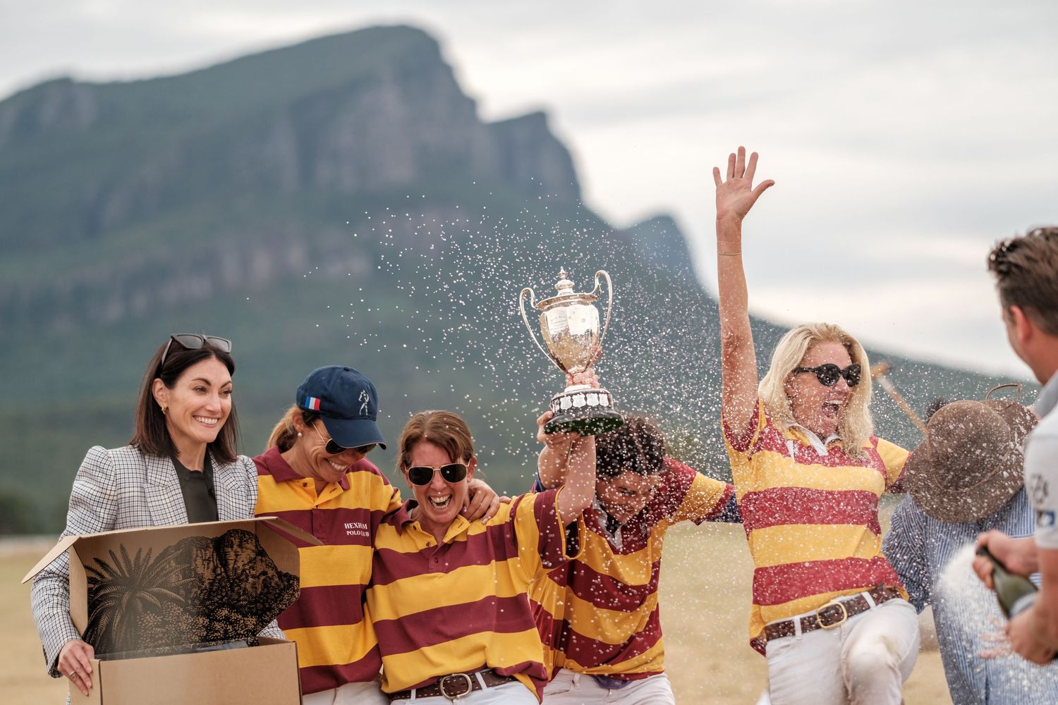Celebratory crowd spraying champagne outdoors with mountain backdrop. Dunkeld Polo winners