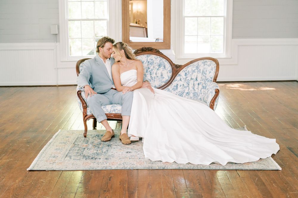 A couple in elegant wedding attire poses gracefully on a white and blue vintage sofa at a historic home wedding venue in Holly Springs NC