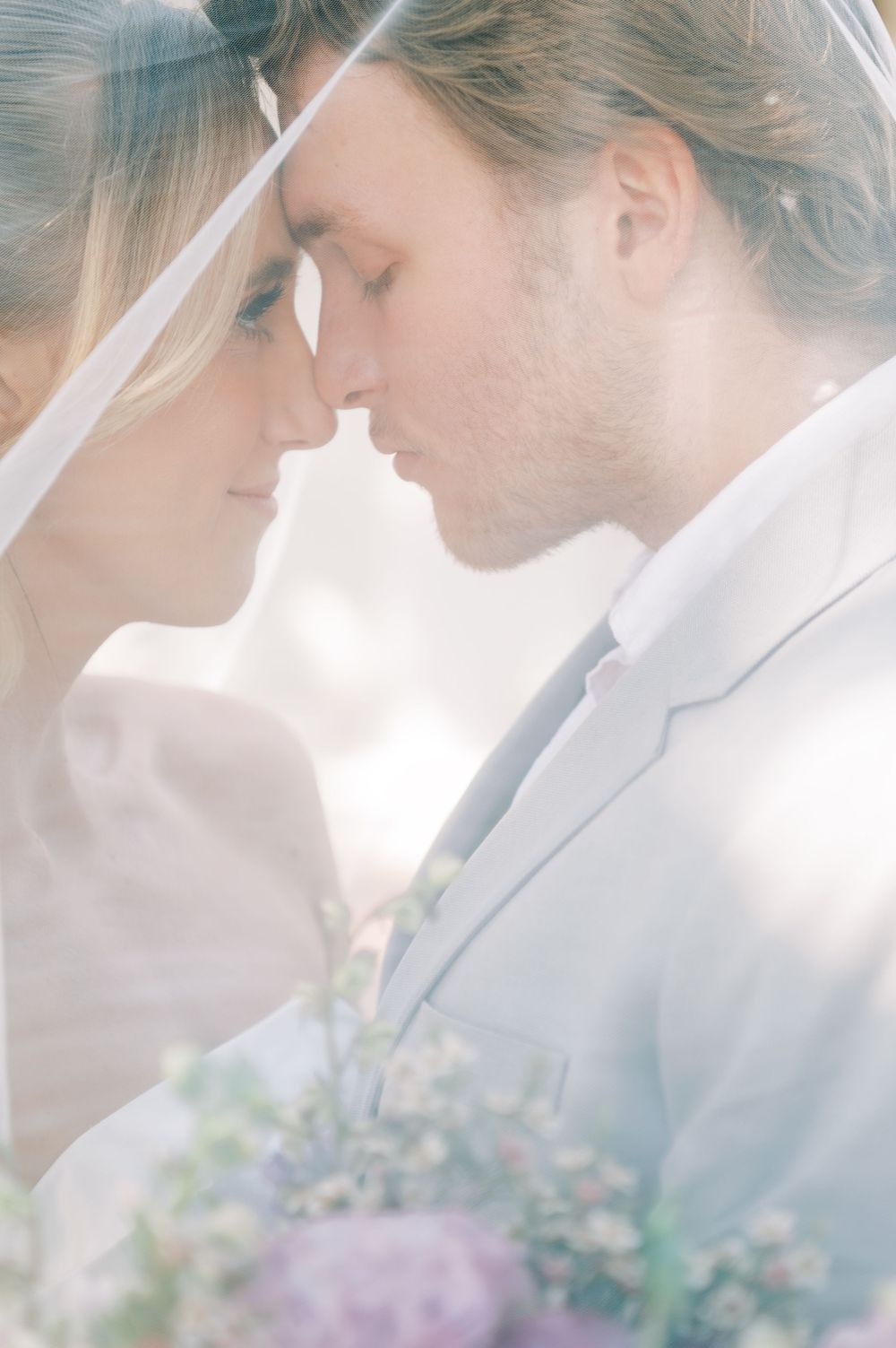 Tender wedding moment captured in soft light with delicate veil and purple floral bouquet.