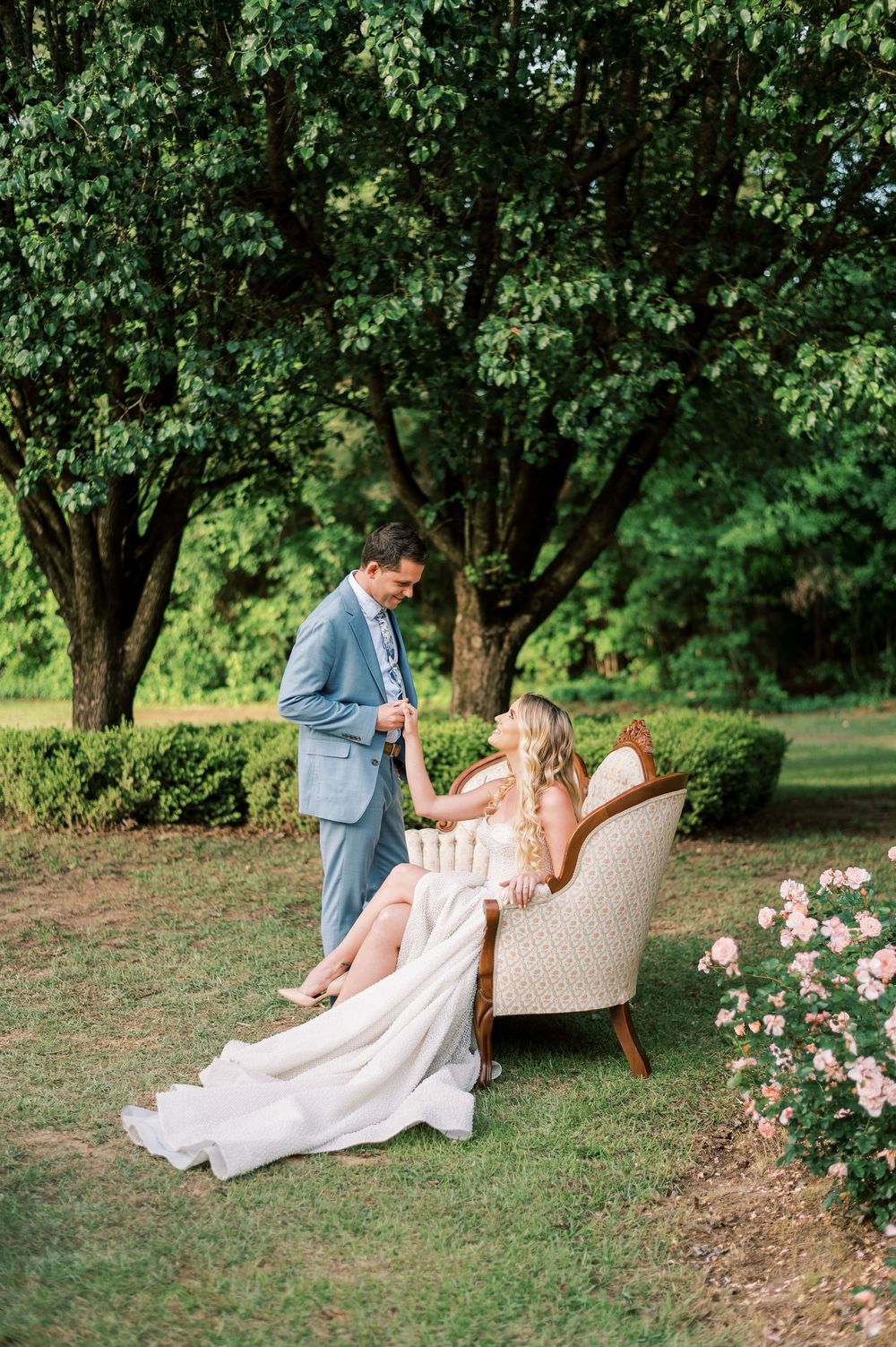 A romantic couple reclines on a vintage settee in a lush garden setting surrounded by blooming flowers and greenery in Fayetteville NC