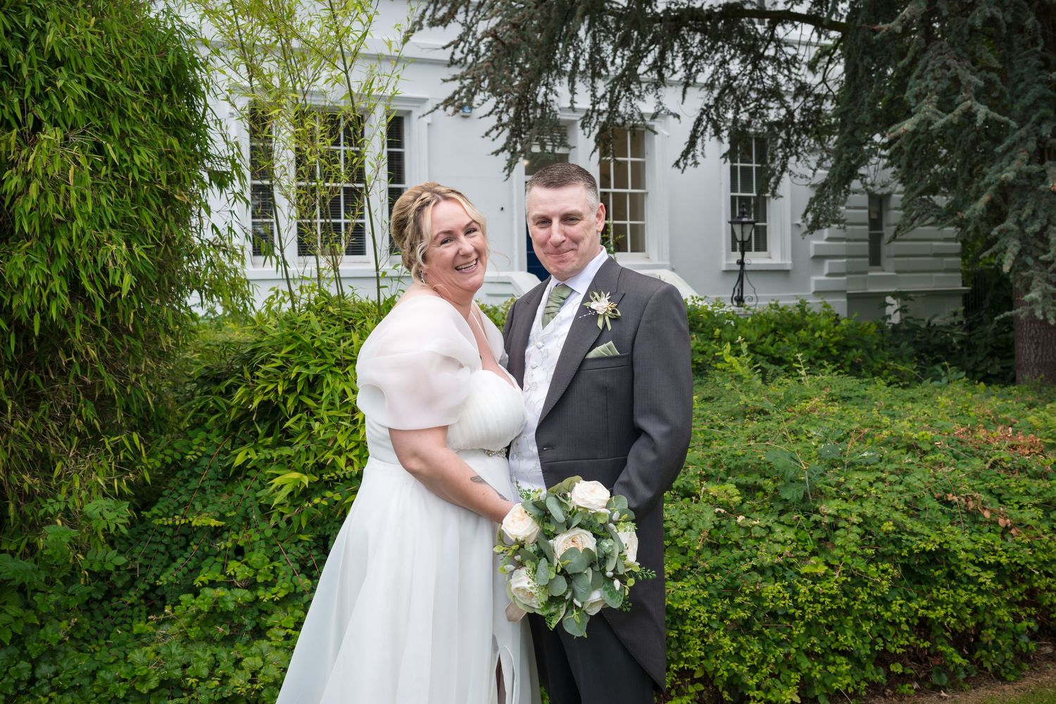 A couple in wedding attire poses together in front of a white building surrounded by lush green foliage.