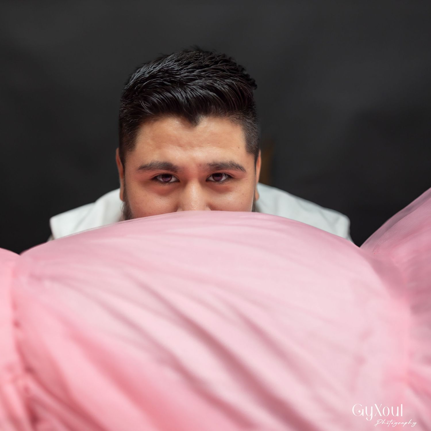 Person peeking over pink pillow or bedding against dark background in playful expression.