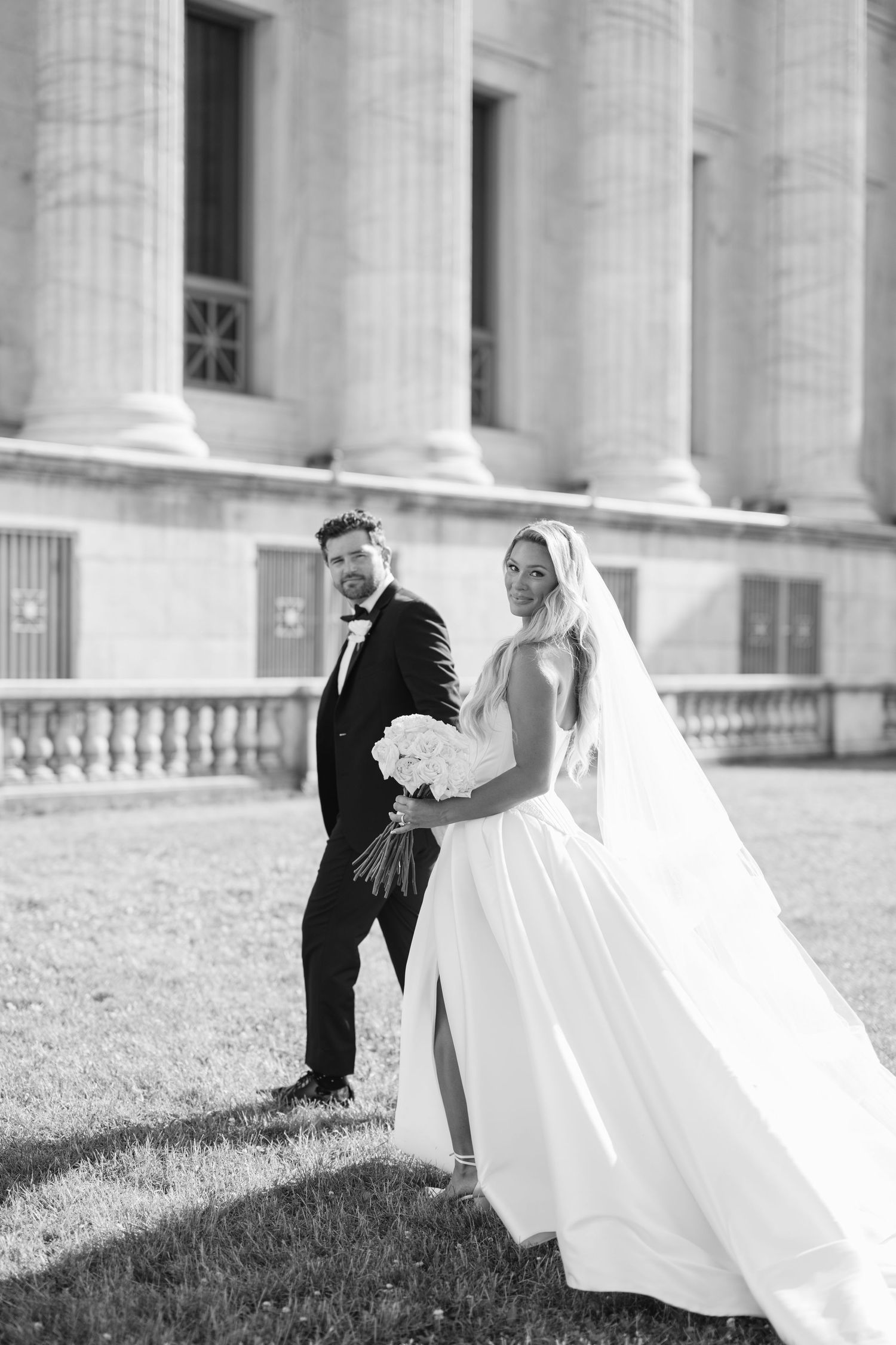 Wedding couple poses elegantly outside grand white columned building with lush green lawn on sunny day.
