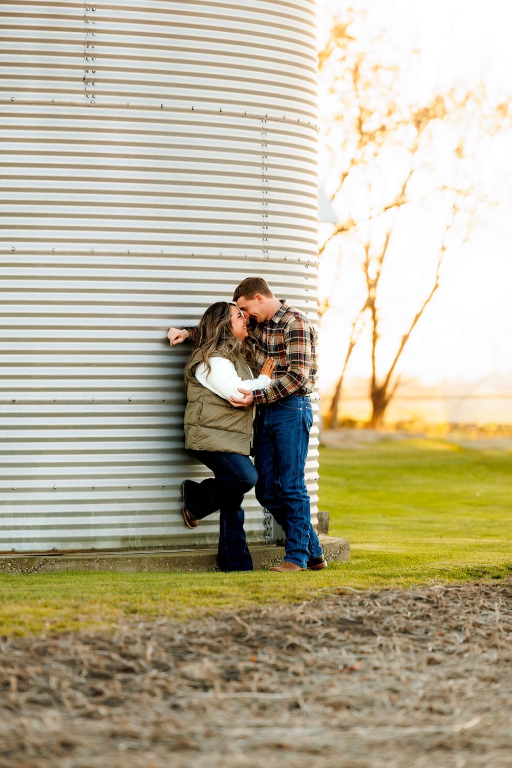 A couple embraces against a large grain silo at sunset in a rural farm setting.