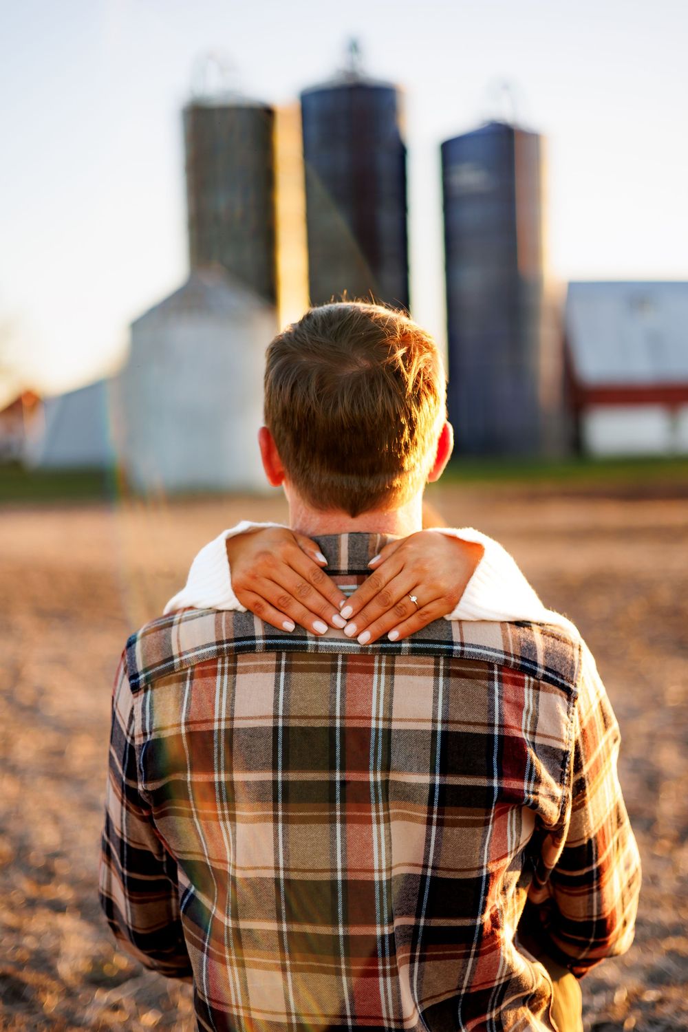 A person in a plaid shirt sits on farmland at sunset with grain silos silhouetted in the background.