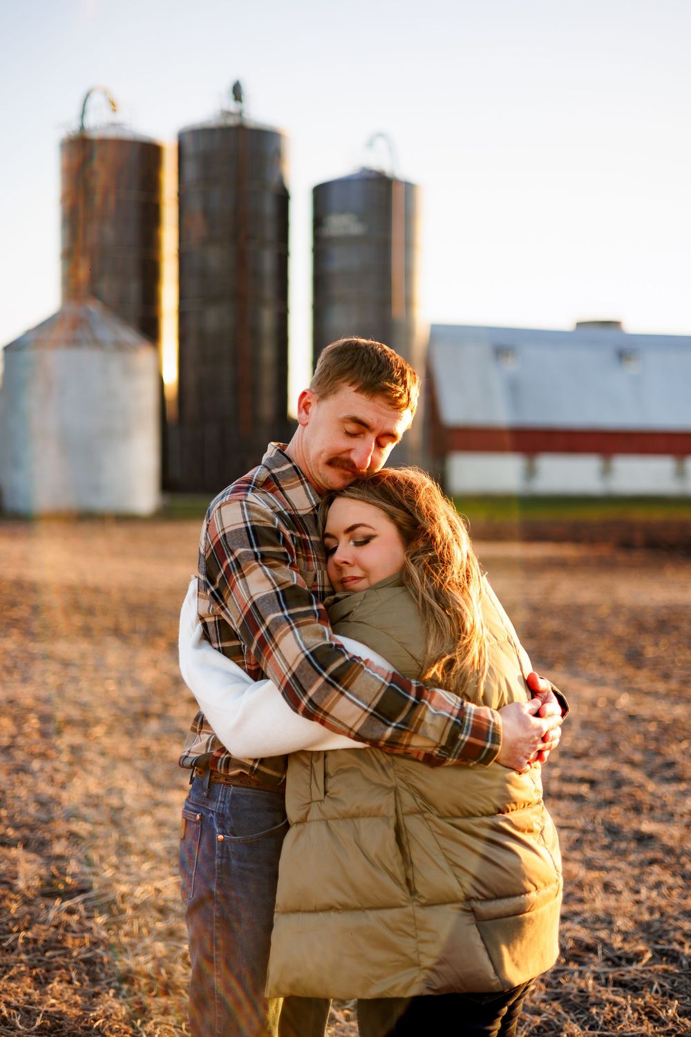 A couple embraces in a romantic moment at sunset on a farm with grain silos in the background.