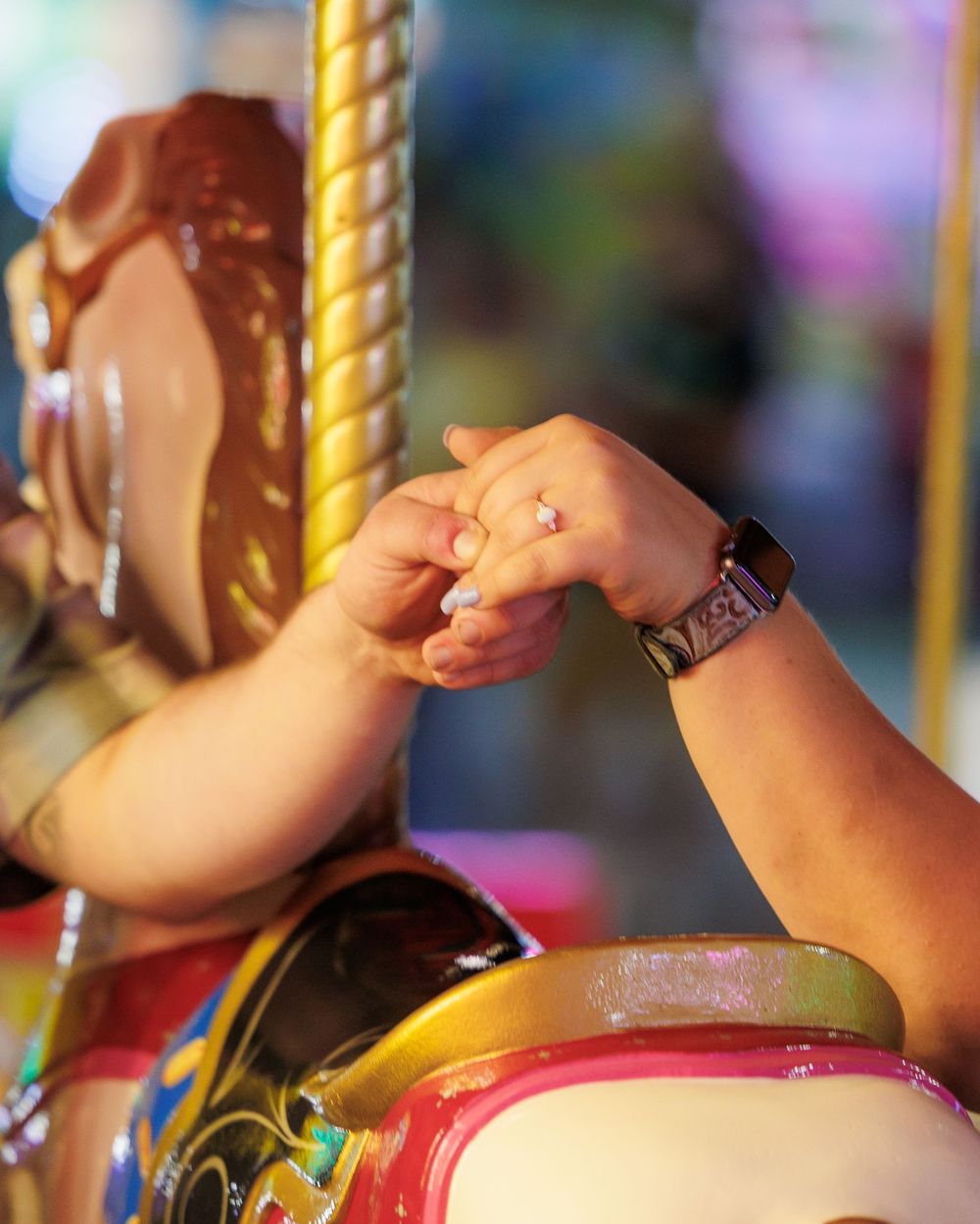 Hands clasp together on a colorful merry-go-round carousel with blurred background lights and decorative elements.