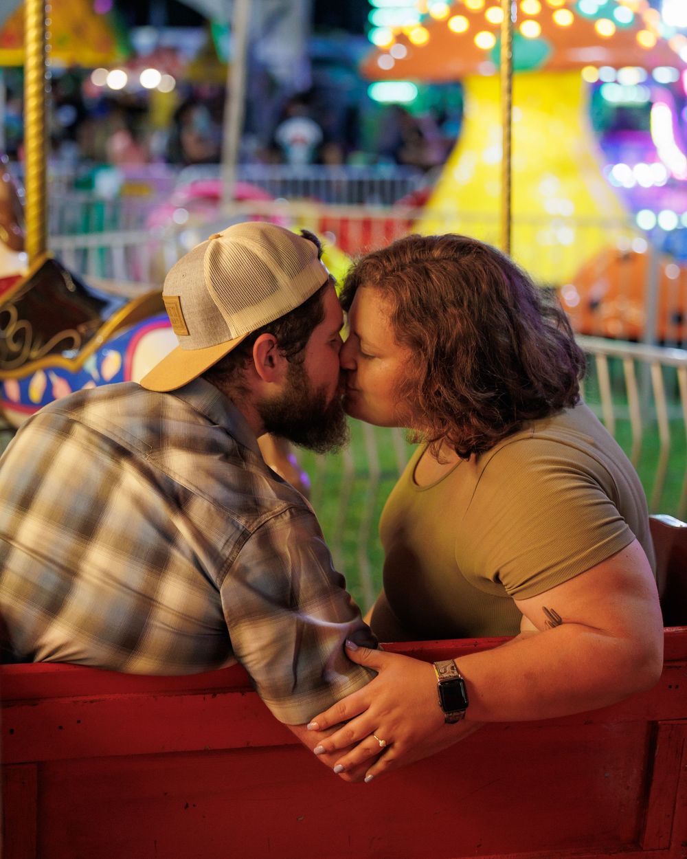 A couple shares a tender kiss while seated at a carnival, with colorful blurred lights and rides in the background.