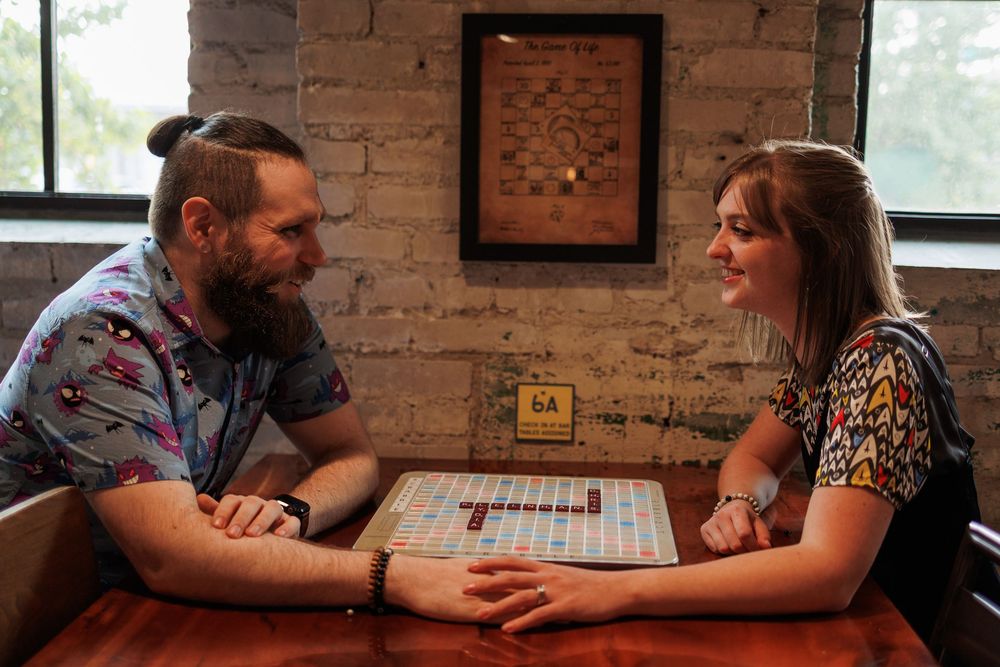 Two people play a board game at a rustic table with exposed brick walls in the background.