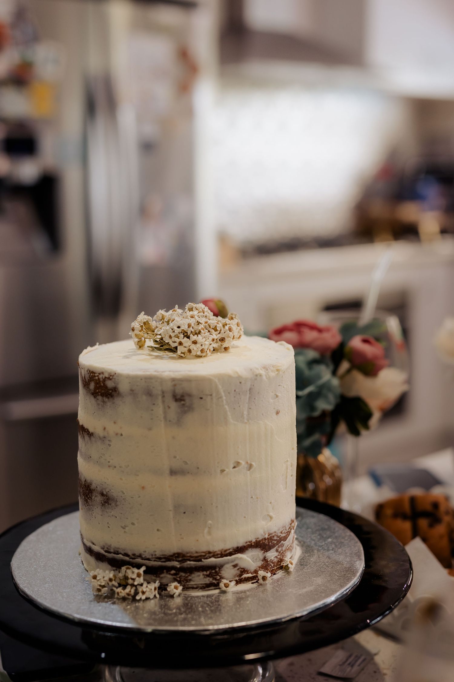 Semi-naked rustic wedding cake with white frosting and dried floral topper on black cake stand in kitchen setting.