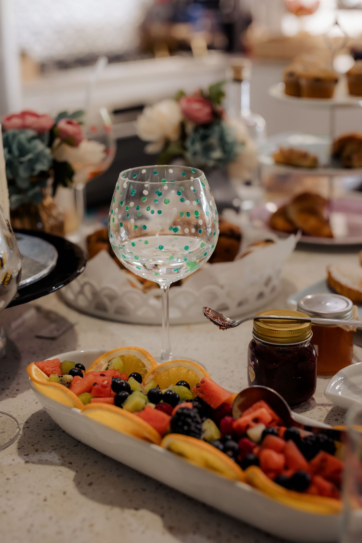 A decorated wine glass sits beside a colorful fruit platter at an elegant table setting with floral decor and mason jars.