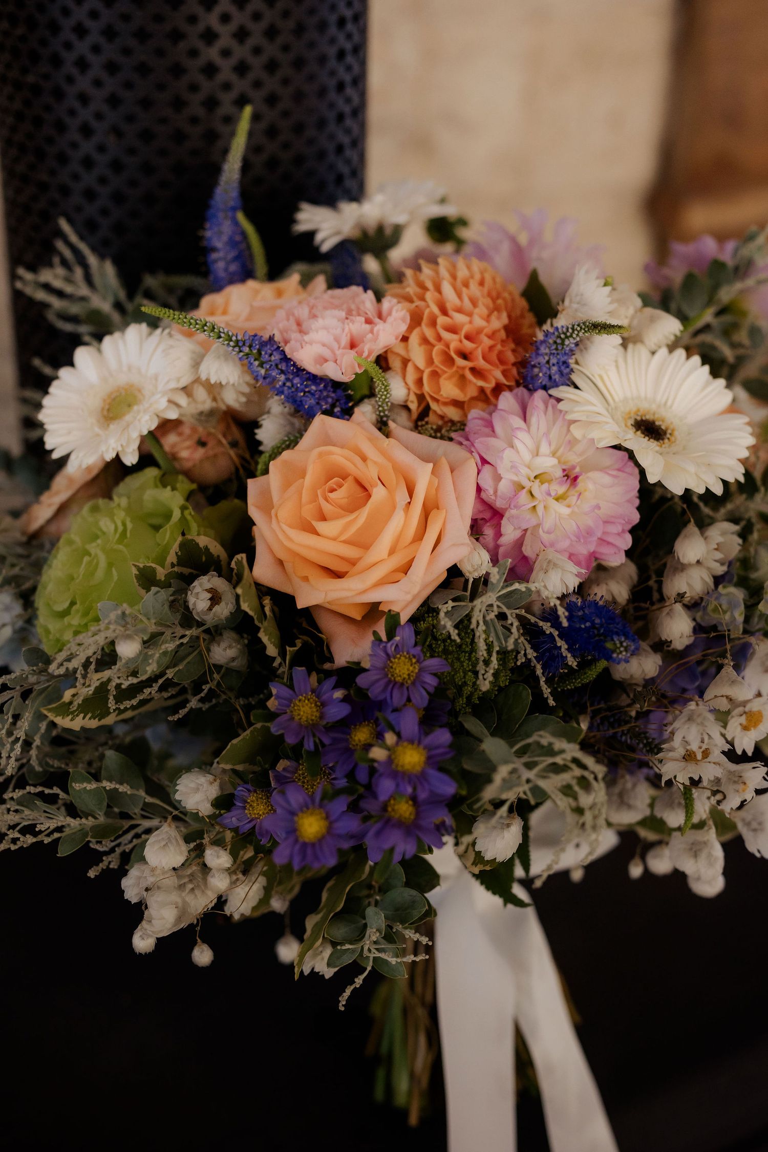 A romantic wedding bouquet with peach roses, white daisies, purple flowers and greenery tied with a white ribbon.