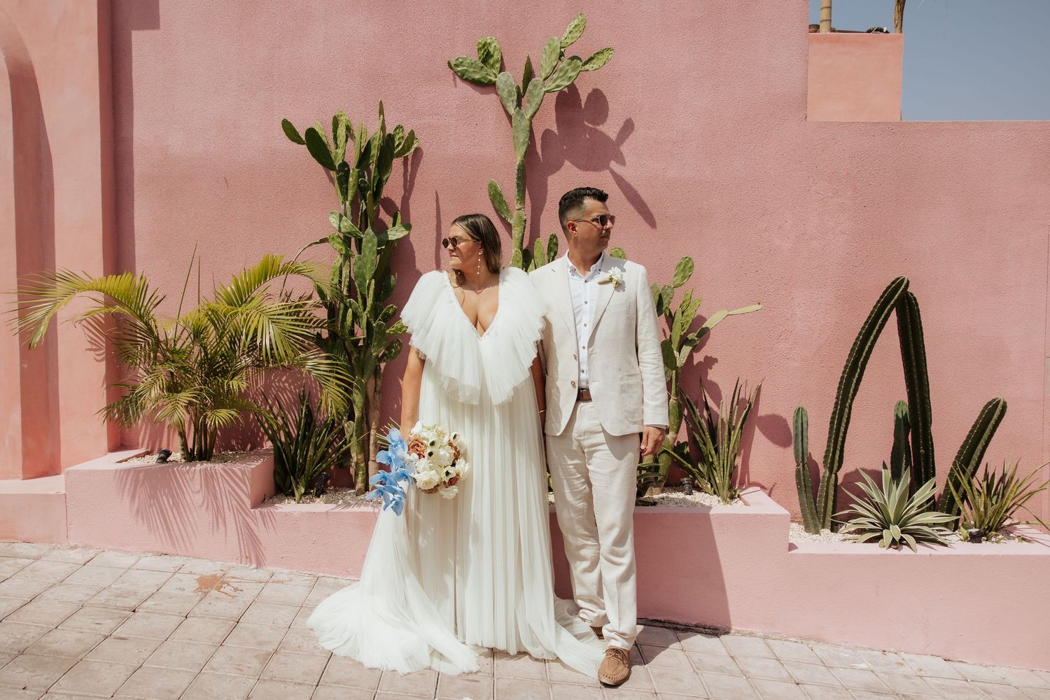 A couple poses together on pink coral-colored steps with Mediterranean architecture and palm trees in the background.