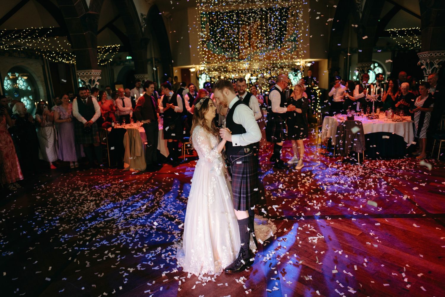 A bride and groom hold hands in a sunlit forest, the groom wearing a traditional Scottish kilt.