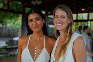 Two women in white dresses smiling warmly while standing together at an outdoor gathering.