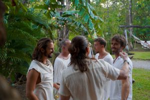 Group of people in white clothing gathered in a lush garden setting having animated conversation.