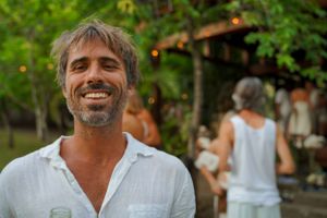 Person in white shirt smiling brightly with tropical garden and gathering in background.