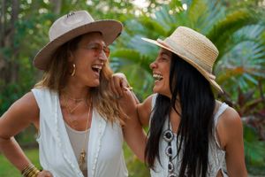 Two friends in white outfits and sun hats sharing a laugh in a tropical garden setting.