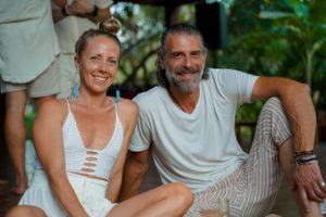 Two people in casual white clothing sitting together outdoors in a tropical setting.