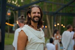 A smiling guest in a sleeveless white top enjoys an outdoor gathering at dusk with string lights in the background.