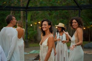 Guests in white attire mingle at an outdoor party with twinkling lights and lush greenery.