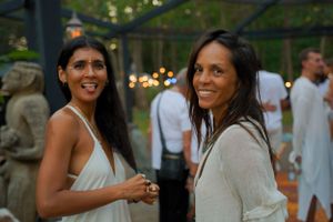 Two guests in white outfits share a moment at a garden party with decorative lighting.
