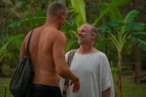 Two guests share a friendly moment at an outdoor event with tropical foliage in the background.