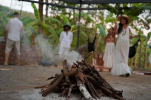 A campfire burns in the foreground with people gathered in white clothing against a jungle backdrop.