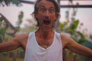 A person in a white tank top expressing extreme excitement with arms outstretched outdoors.